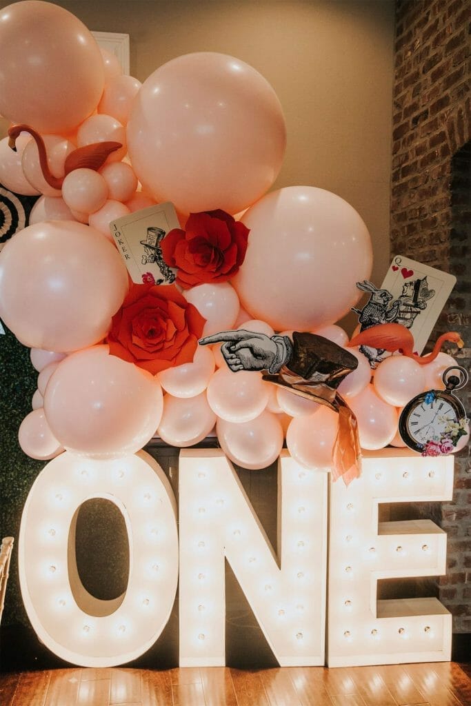 Lighted marquee letters spell “ONE” topped with pink balloons, giant red roses, and oversized playing cards for an Alice in Wonderland birthday party.